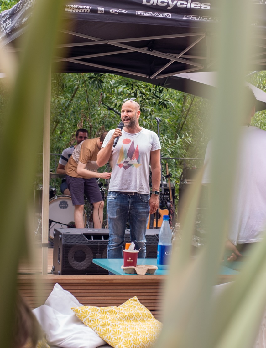 Jan Schmirmund spricht mit Mikrofon auf einer Bühne im Freien, hinter ihm eine Band, vorn unscharfe Blätter
