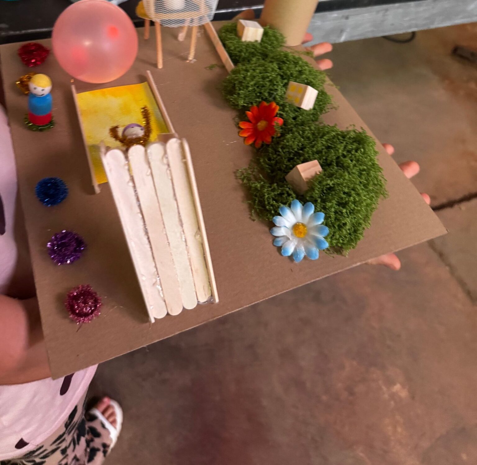 Eine Hand hält ein selbst gebautes Modell eines Spielplatzes mit Rutsche, Moos und Blumen
