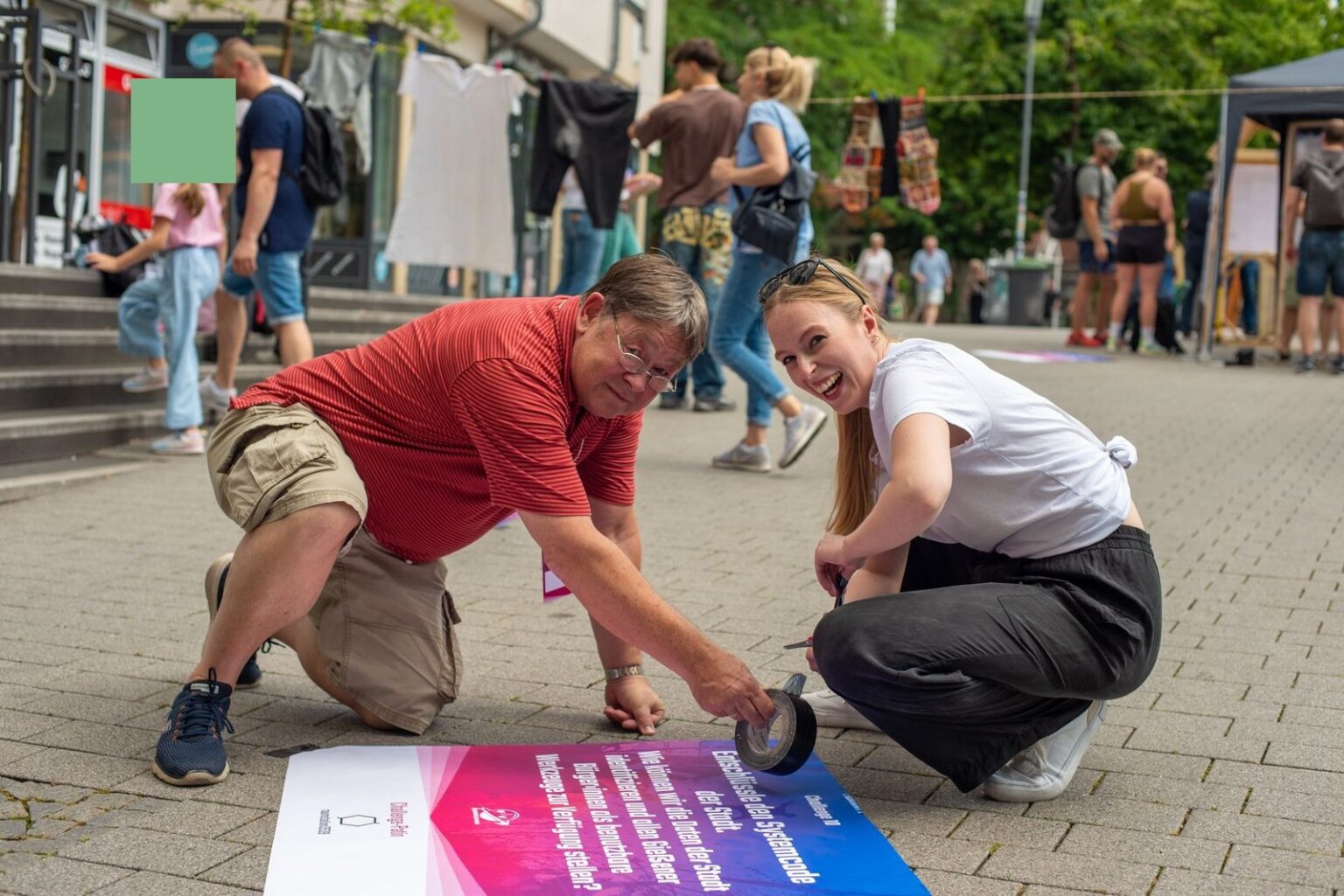 Zwei Menschen kleben in einer Fußgängerzone lachend ein Plakat auf den Boden