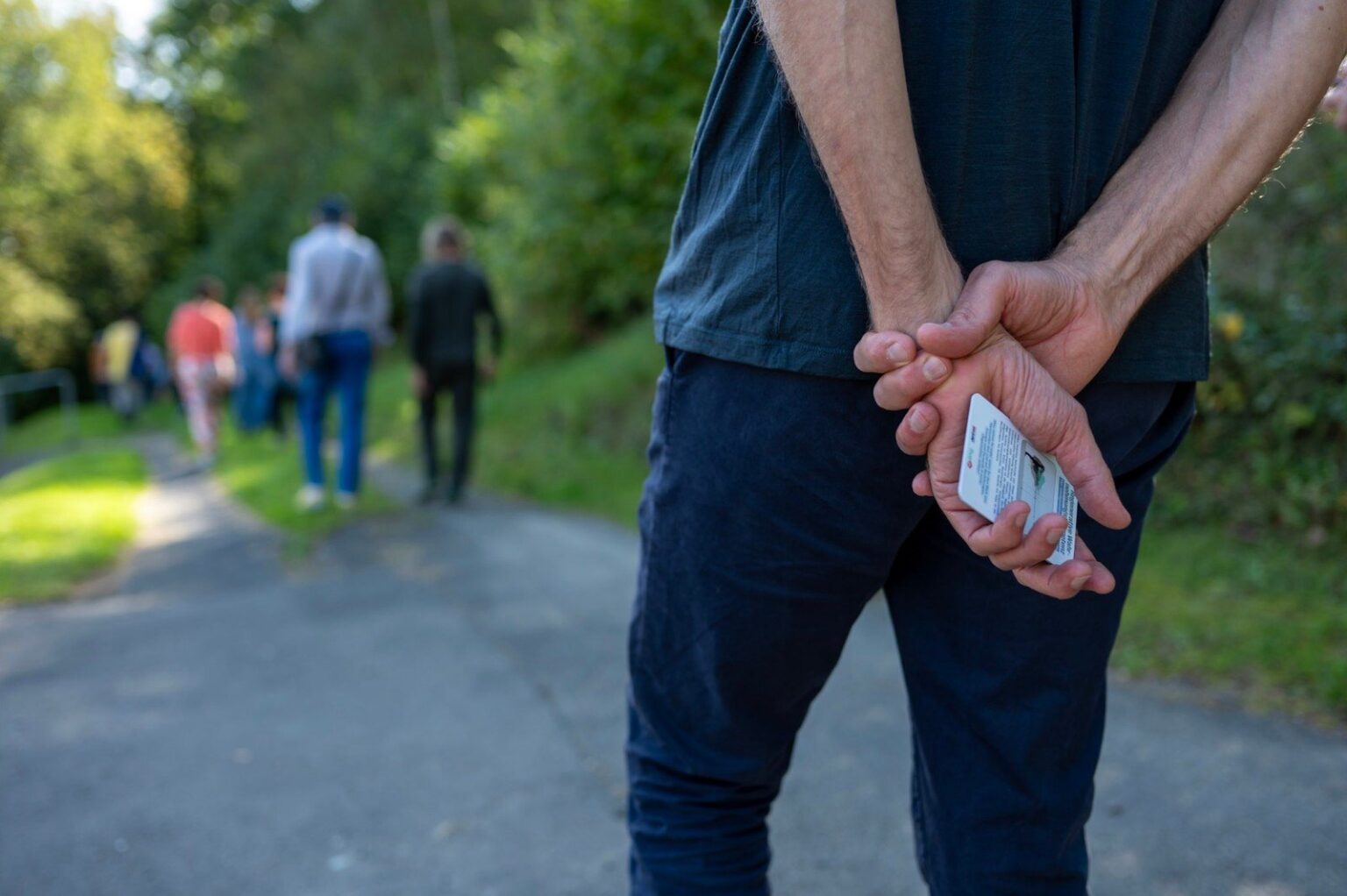 Auf einem Waldweg hält jemand Gesprächskarten hinter dem Rücken, weiter vorne geht eine Gruppe spazieren